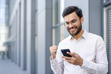 A cheerful man in glasses celebrates good news on his phone, smiling brightly outdoors near a...