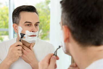 Handsome man shaving with razor near mirror at home