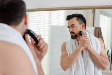Handsome man shaving with trimmer near mirror in bathroom