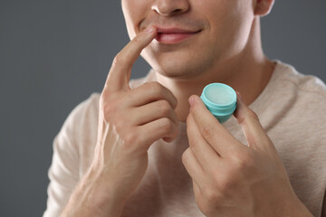 Young man applying lip balm onto his lips on grey background, closeup