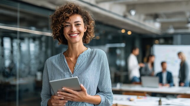 Professional smiling woman holding digital tablet in modern office environment with colleagues.