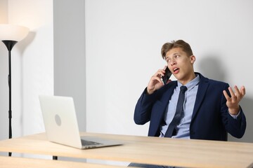 Young businessman with laptop complaining to someone via phone at wooden table indoors