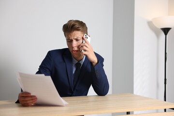 Young businessman with papers complaining to someone via phone at wooden table indoors