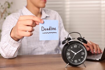 Man holding paper note with word Deadline while working and alarm clock on wooden table, selective...