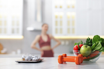 Slim woman in sportswear posing indoors, focus on kitchen scale, healthy food, dumbbells and...
