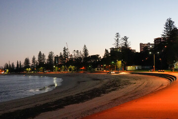 Twilight shoreline view of Redcliffe beach promenade in Queensland, Australia with silhouetted pine trees, lit footpath, city buildings, and calm waves under a clear dusk sky.