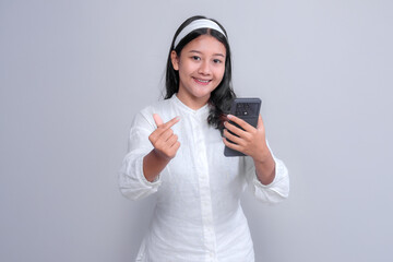 Asian woman with long black hair wearing a white shirt and headband, smiling while holding a smartphone and making a finger heart gesture on a plain background.