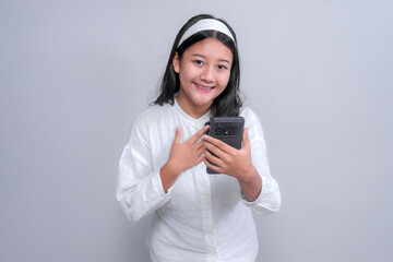Portrait of a cheerful young Asian woman wearing a white headband and shirt, smiling warmly while holding a smartphone and touching her chest in a thankful gesture, isolated on a clean