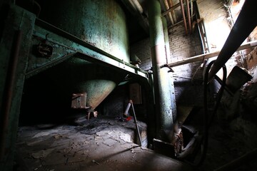 Rusted green tank beneath corroded pipes in a dimly lit corner of an abandoned factory