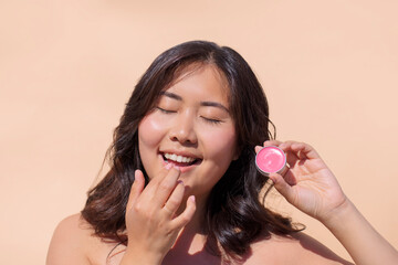 Asian woman applying lip balm with a joyful expression, holding a small container of pink product...