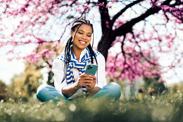 Young woman sitting outdoors beneath blooming trees enjoying her smartphone on a sunny spring day in the park