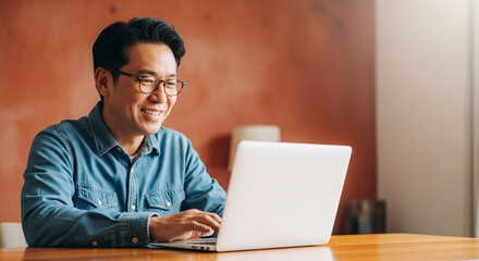 Man with glasses smiling while using a laptop at a wooden table in a room with a warm wall color