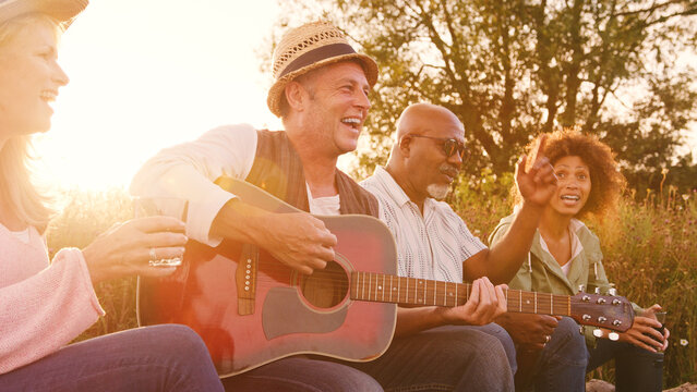Group Of Mature Friends Sitting Around Fire As They Drink And Sing Songs At Outdoor Campsite