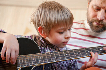 Handsome dad teaches his cute son to play the guitar sitting on sofa in the living room at home....