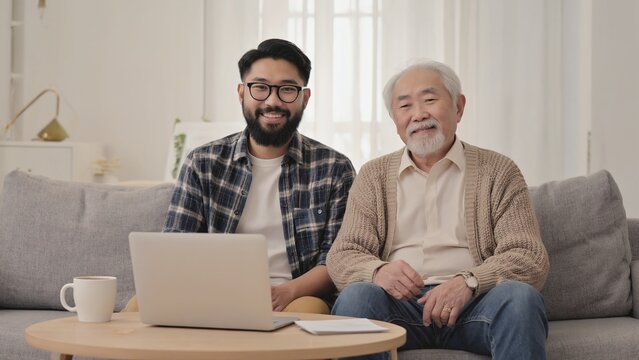 A young Asian man with black hair and glasses sits next to a senior Asian man with gray hair. They are smiling while using a laptop in a cozy living room.