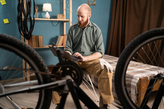 Bearded male mechanic or worker with laptop analyzing broken bike working at home. replacement parts for bicycle online - Powered by Adobe