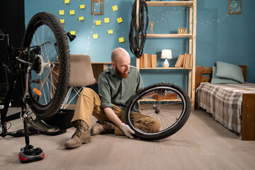 Bearded man sitting on floor holding bicycle wheel repairing bike at home. Bicycle repair and...