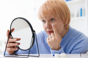 Senior woman putting on contact lenses at table near mirror in ophthalmologist office