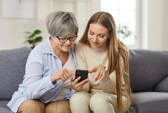 Happy mother and adult daughter spending leisure time together at home, using a mobile phone for online entertainment in internet. Family connection through technology and digital bonding.