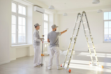 Two professional contractors in work uniforms standing by ladder in bright empty room, discussing...