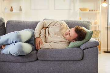 Sick young man lying on sofa, holding his stomach with pained expression. Unhealthy male person...