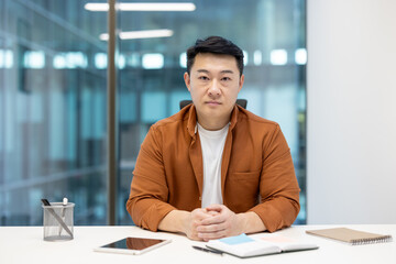 A focused Asian man sits at a desk, looking directly at the camera in a modern office setting,...