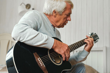 Obraz premium Senior man is playing guitar. Elderly male sitting at sofa and play music. Portrait of a gray haired mature 60s person with acoustic guitar, he is learning to play. Enjoying retirement life at home.