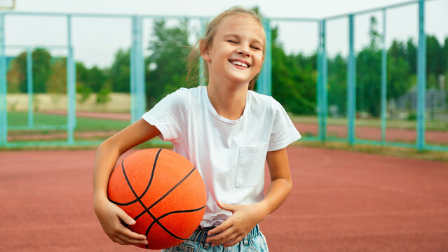 Cheerful child girl basketball player holding game ball standing on the basketball court outdoors