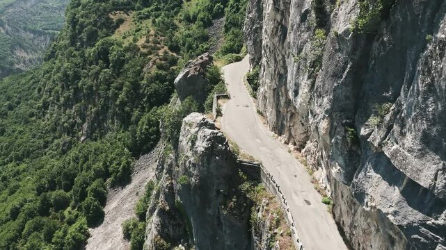 Mountain road carved along steep cliffside, surrounded by dense green forest and rocky terrain, dramatic drop on one side, drone shot.