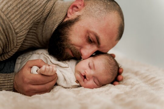 A tender moment between a father and his newborn baby, both lying on a soft surface. The father is gently kissing the baby's forehead, showcasing love and affection.