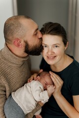 couple with a newborn baby. The father is kissing the mother's forehead while she holds the sleeping baby close. The scene conveys warmth and family love.