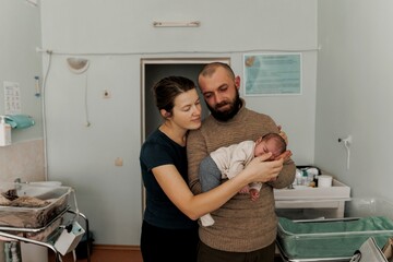 A couple lovingly holds their newborn baby in a cozy indoor setting. The mother gazes at the baby...