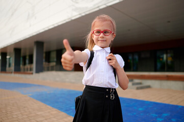 young schoolgirl with backpack gives thumbs-up in front of modern school building, wearing red...