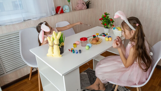 Two cute sisters painting eggs and preparing for Easter. Girls wearing bunny ears or rabbit ears on head and holding painted easter eggs (dyed eggs) in hands.