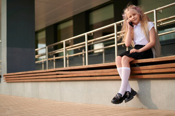 Obraz premium schoolgirl with smartphone sitting on bench outdoors. young student in uniform chatting or calling, enjoying sunny day. back to school concept, education lifestyle theme