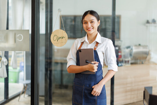 Portrait of happy asian woman standing and holding digital tablet at open sign of her store.  Successful small business owner in casual wearing blue apron standing at entrance cafe.
