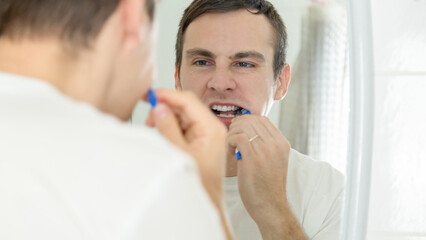 Young man brushing teeth with toothbrush while standing before mirror at home in the bathroom
