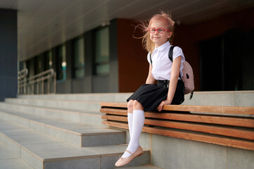 schoolgirl with glasses sits on bench near school building, wearing uniform and backpack. ready for...