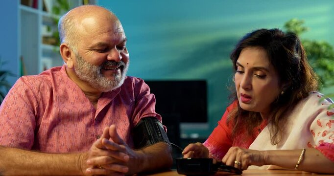 Senior couple measuring blood pressure at home using digital BP monitor sitting together at the table, Indian retired family happy with healthy readings, wellness, aging, healthcare lifestyle concept
