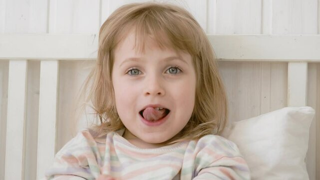 4 years girl joking, sticking tongue out with silly expression. Child making face. Funny foolishness portrait of little kid doing crazy comical gesture among a white wall. Closeup face. Low contrast