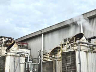 Industrial cooling tower system emitting vapor under blue sky. Heavy-duty piping, valves, and heat exchangers in a manufacturing or refinery plant exterior.