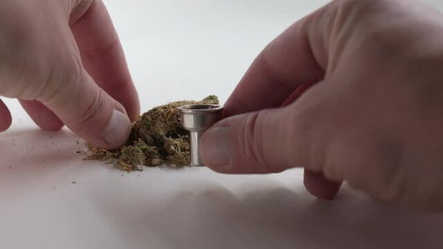 Hands of anonymous person loading marijuana into pipe from white surface, preparing for use in a focused and careful manner