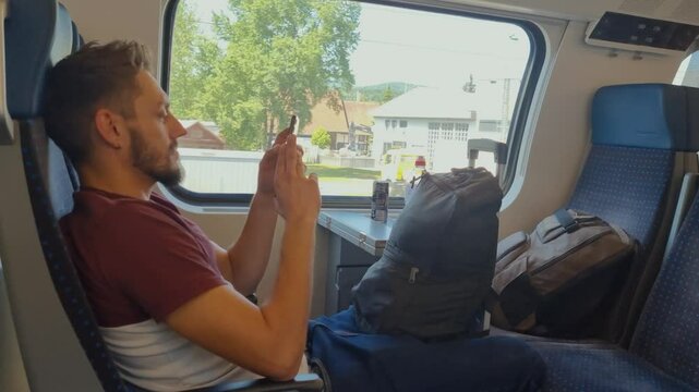 A man sits relaxed on a Swiss train with his luggage, scrolling on his phone as Swiss buildings and a spring landscape pass by outside the window