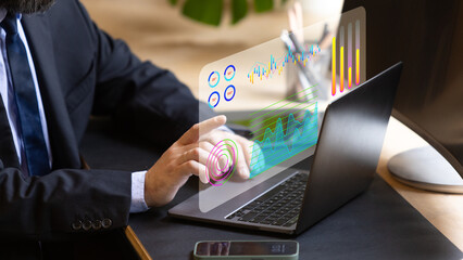 Close-up of suited man's hand operating a digital transparent screen with graphs and analytics on a laptop. Futuristic office setting with advanced technology in business operations.