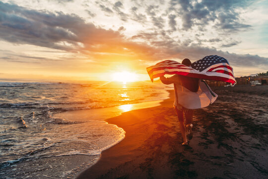 Young woman holding national American flag walking ocean beach. America Independence Day concept - Powered by Adobe