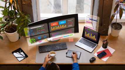 Top view of creative digital analyst working at a desk with curved monitor, laptop, and holographic infographics. Concept of smart work environments, creative economy workspace