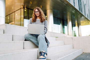 Stylish woman with laptop sitting on steps against modern building background. Business woman in glasses working outdoors on laptop. Freelance, business and break concept.