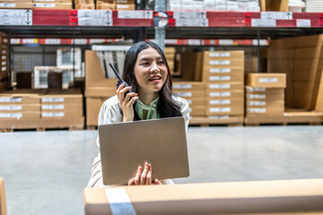 lively warehouse, dedicated young asian woman meticulously checks inventory details on her laptop while engaged in a conversation for radio communication, ensuring smooth operations and accuracy