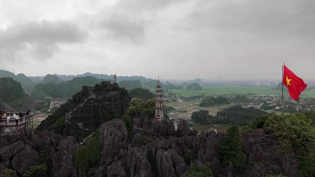 Aerial drone footage of a Vietnamese flag waving in the wind on Hang M&uacute;a viewpoint, with a stone pagoda and lush green landscapes in Ninh Binh, Vietnam