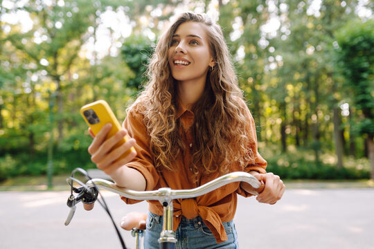 Nice woman with phone and bicycle enjoying time outdoors. Beautiful woman riding bicycle and texting on phone in sunny park. Weekend concept. Active lifestyle.
